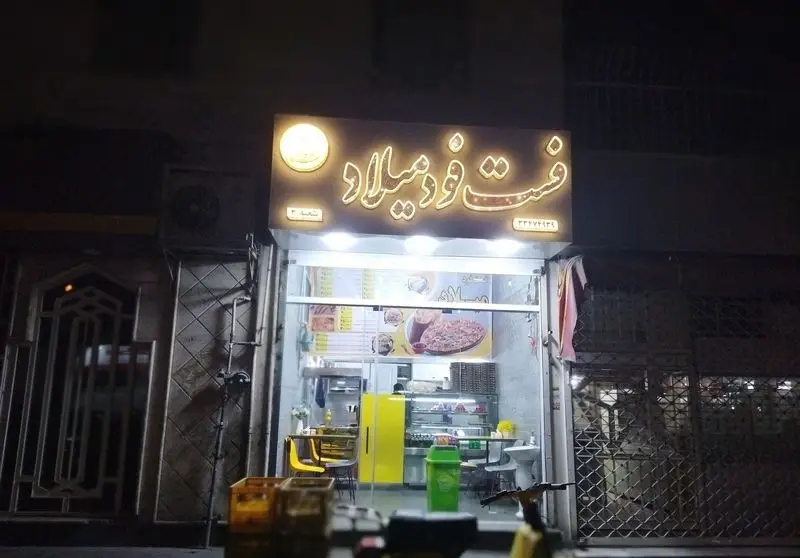 فست فود پیتزا و ساندویچ میلاد 2 در شب، با تابلوی روشن و منوی متنوعی از پیتزاها و ساندویچ‌های خوشمزه آماده پذیرایی.