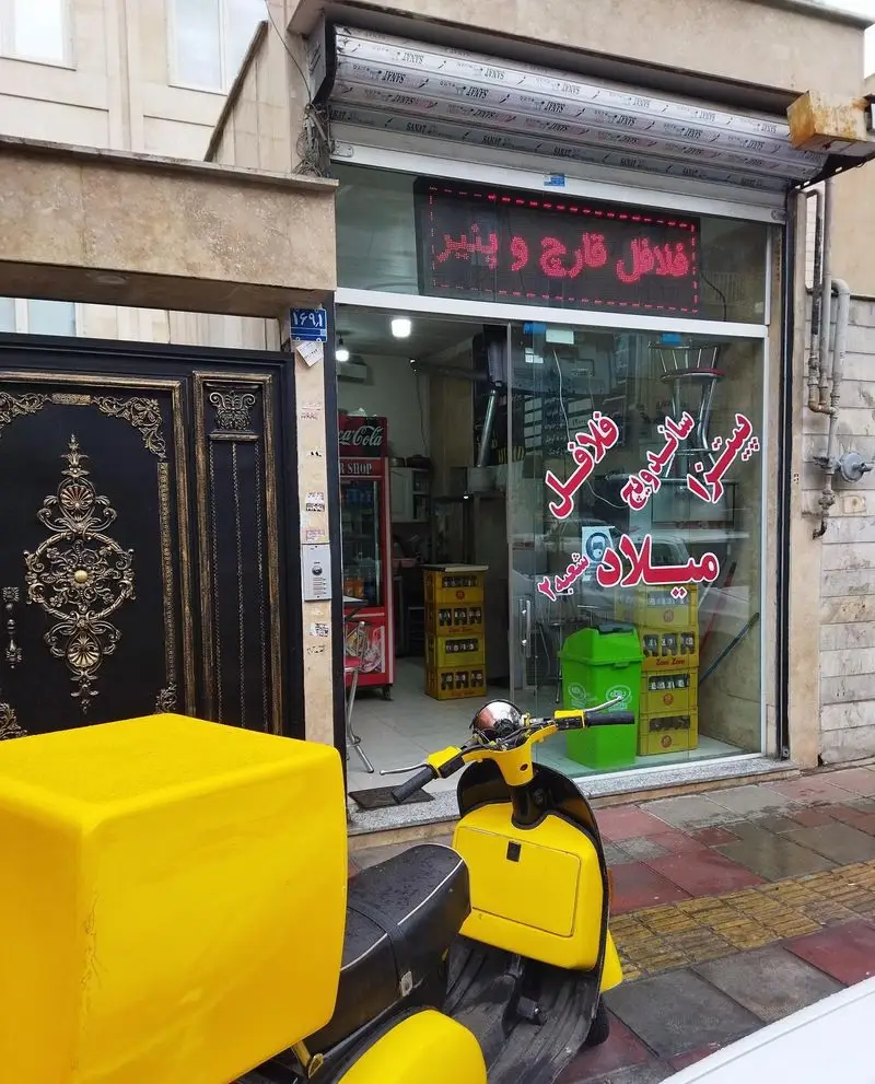 تصویری از نمای بیرونی مغازه پیتزا ساندویچ میلاد 2 با تابلوی فلافل قارچ و پنیر و اسکوتر دلیوری زرد در پیاده رو. غذای آماده میلاد.