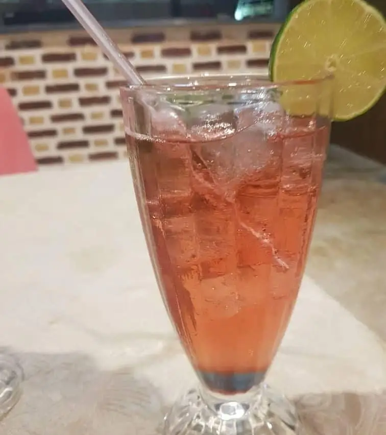 یک لیوان بلند نوشیدنی خوشرنگ صورتی با یخ و لیموترش، آماده سرو در محیط آرام و زیبای رستوران پرشین گلف، انتخابی دلچسب.