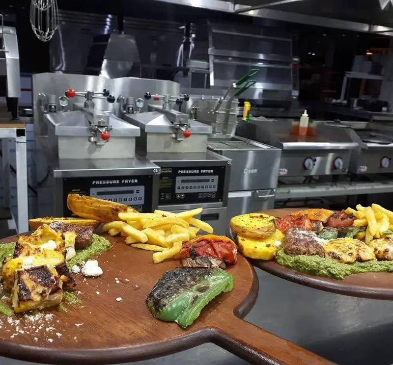 سینی های چوبی پر از غذاهای گریل شده لذیذ (کباب، جوجه، سبزیجات) و سیب زمینی سرخ کرده در آشپزخانه مدرن رستوران پرشین گلف.