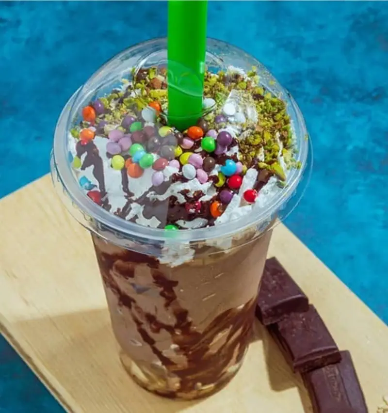 میلکشیک شکلاتی آبمیوه و بستنی عارف با تزئین خامه، اسمارتیزهای رنگی، پودر پسته و سس شکلات، همراه با تکه های شکلات تلخ