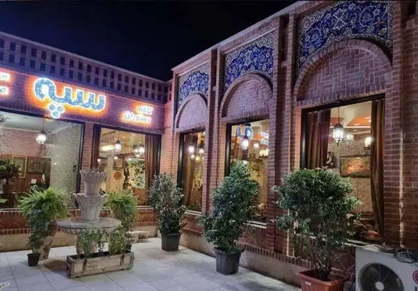 نمای شب کافه و رستوران سنتی سپه با معماری آجری، کاشی‌کاری سنتی آبی، آبنما، گلدان‌ها و فضایی دلنشین و دنج.