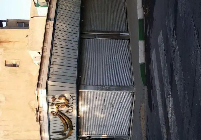 تصویری از نمای بیرونی قنادی بهارلو با کرکره‌های فلزی بسته، تابلوی طلایی زیبا و پیاده‌رو کنار خیابان در یک روز روشن.