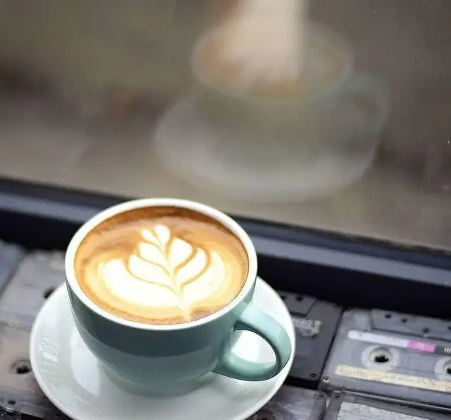 فنجانی آبی از لاته آرت با طرح برگ زیبا، در کافه رستوران پلاک ۵. بازتاب تصویر در شیشه و نوارهای کاست قدیمی در کنار آن.