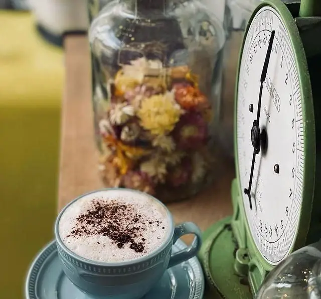 کاپوچینو یا لاته با پودر کاکائو در فنجان آبی روشن در فضای دنج کافه رستوران پلاک ۵، کنار شیشه گل خشک و ترازوی قدیمی.