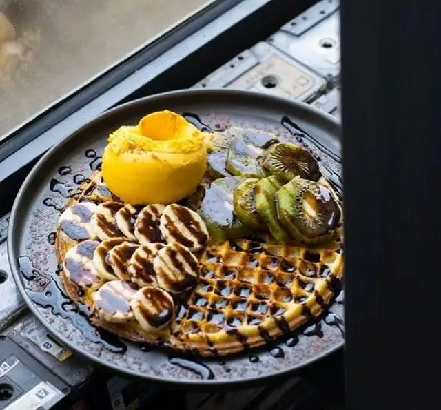 وافل طلایی با موز و کیوی، سس شکلات و اسکوپ بستنی میوه‌ای، تصویری از منوی جذاب کافه رستوران پلاک ٥ برای لحظات شیرین.