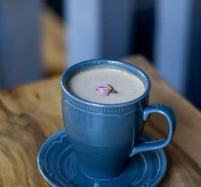 فنجان آبی رنگ نوشیدنی گرم و معطر با غنچه رز صورتی زیبا بر روی آن، در محیط دنج کافه رستوران پلاک ٥.