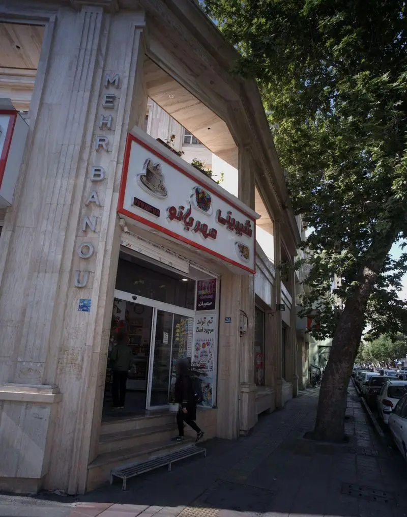 عکس نمای خیابانی شیرینی فروشی مهر بانو در تهران، با ورودی مغازه، تابلو، پیاده‌رو، درخت و صف خودروها در یک روز روشن.