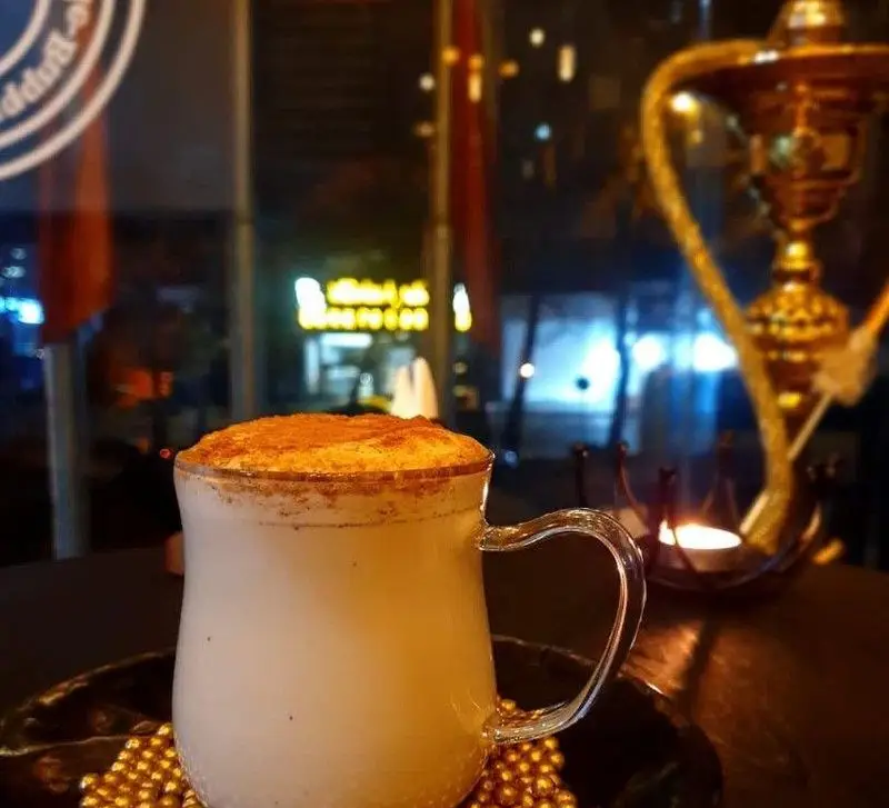 نوشیدنی گرم و دلپذیر لاته یا قهوه در فنجان شیشه‌ای، با تزیین دارچین روی میز کافه ویترای در فضای شبانه و دنج.