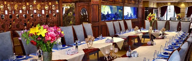 تصویری از فضای داخلی بزرگ و مجلل کافه رستوران ارکیده شعبه آرژانتین با میزهای چیده شده، دکوراسیون چوبی و آکواریوم دیواری.