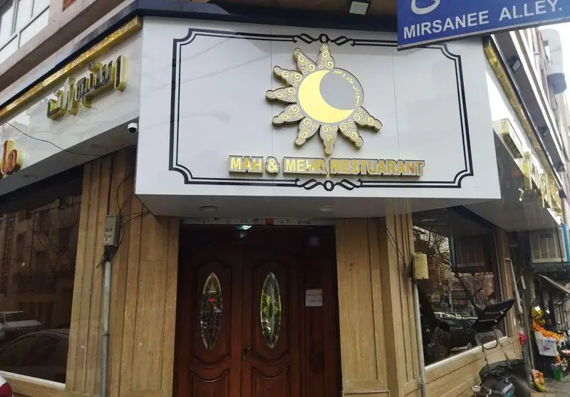 نمای بیرونی رستوران ماه و مهر با تابلوی اصلی، شامل لوگوی ماه و خورشید طلایی، ورودی چوبی شیک و موقعیت آن در کوچه میرثانی.