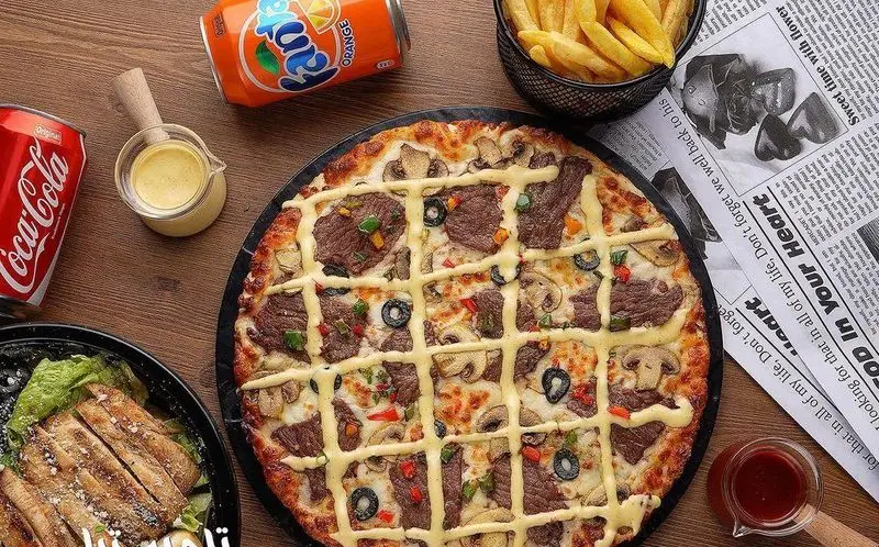 تصویری جذاب از میز پر از غذاهای لذیذ در فست فود تله پیتزا: پیتزای استیک و قارچ، سیب زمینی سرخ کرده، سالاد سزار مرغ و نوشابه‌ها.