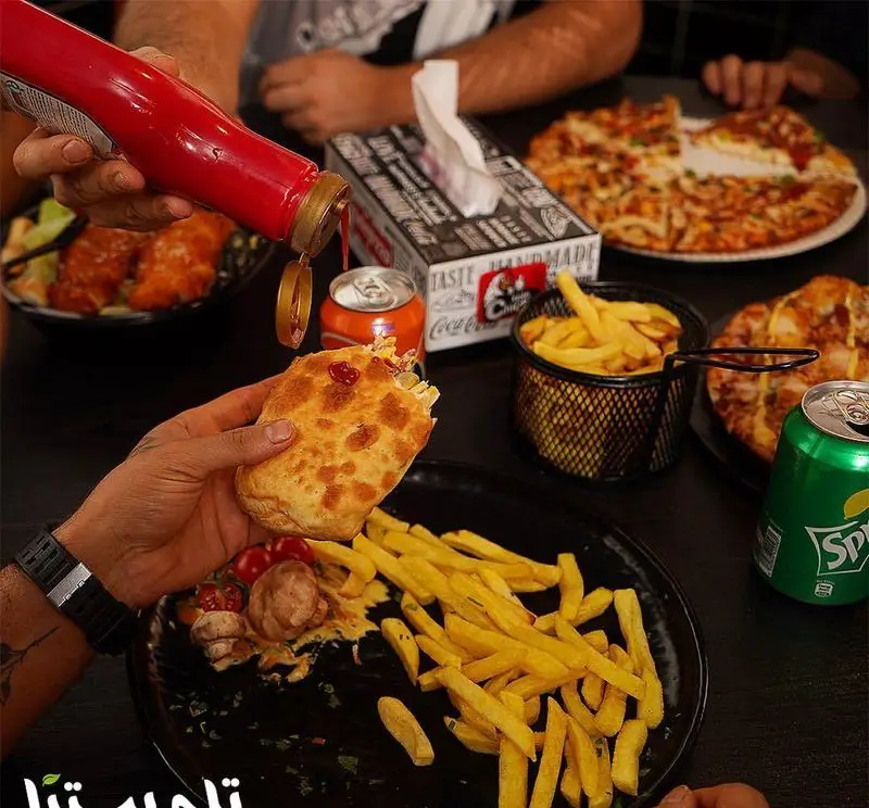 تصویری از محیط شلوغ و لذت‌بخش فست فود تله پیتزا با پیتزاهای داغ، سیب زمینی سرخ کرده و نوشیدنی‌ها، همراه با افزودن سس کچاپ.