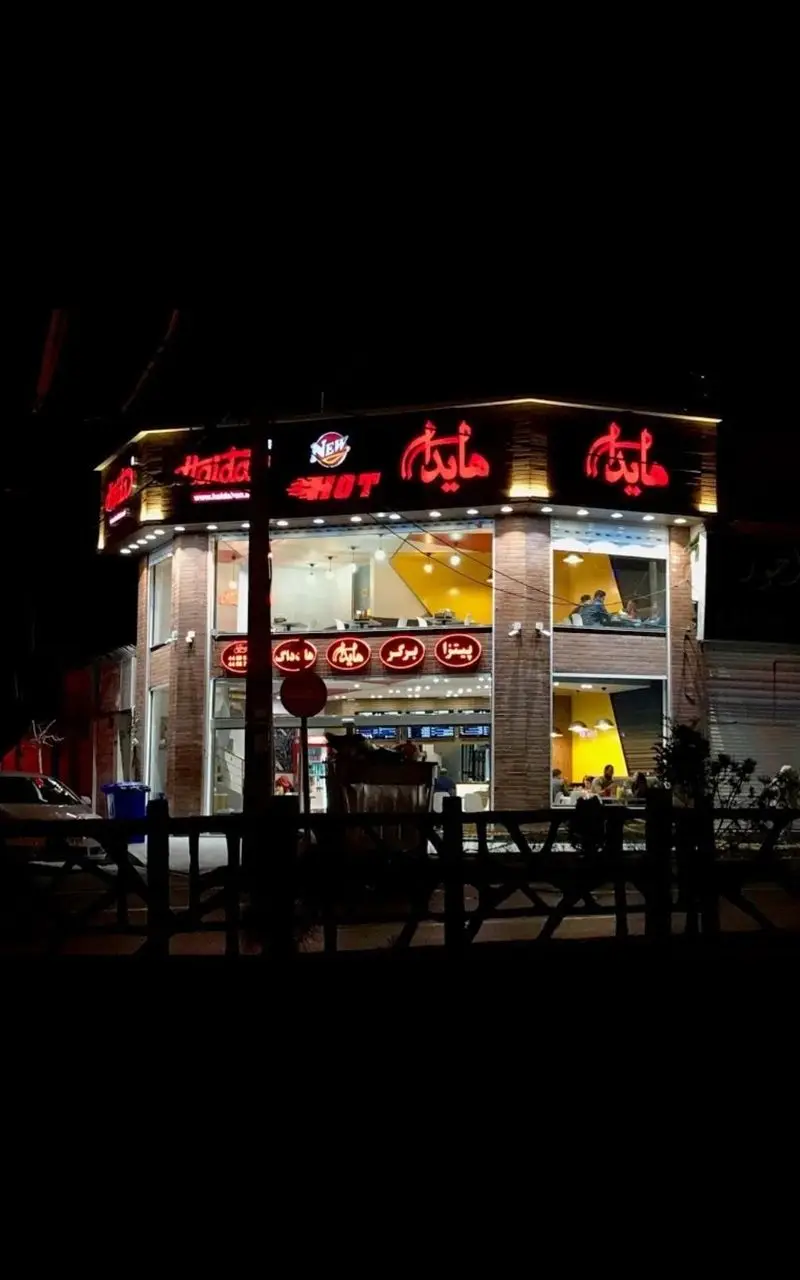 نمای شبانه فست فود هایدا با تابلوهای نئونی درخشان، ساختمان مدرن و مشتریان در حال لذت بردن از پیتزا و برگر.