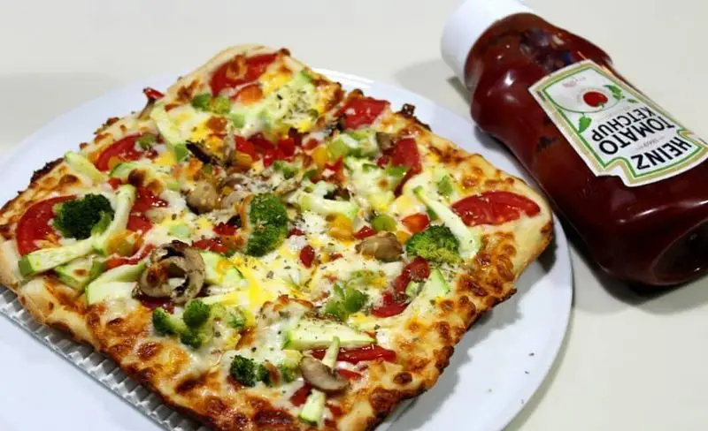 پیتزا سبزیجات مربع رستوران ایتالیایی رو ب رو با بروکلی، فلفل و گوجه، آماده سرو همراه سس کچاپ هاینز، غذایی لذیذ.