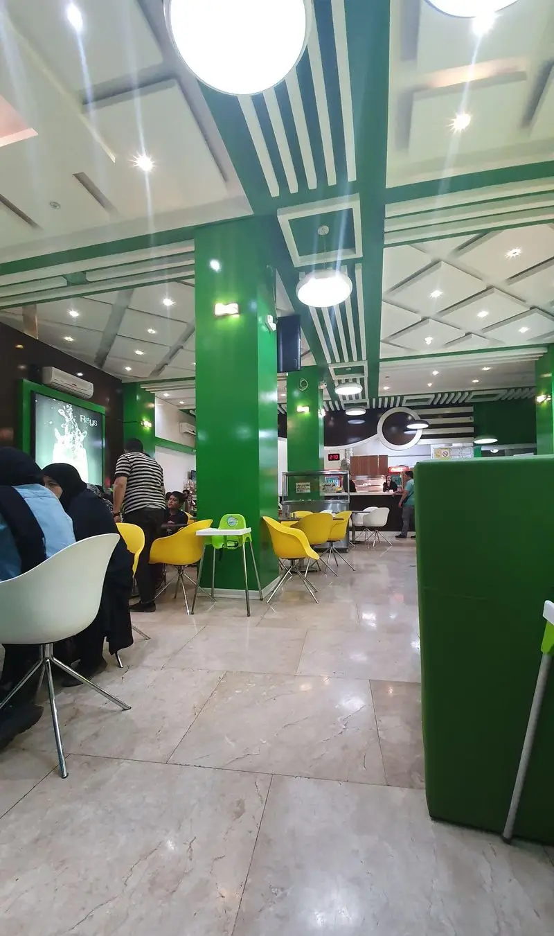 نمای داخلی فست فود صدرا، با طراحی مدرن شامل ستون‌های سبز و سقف راه راه سفید و سبز، صندلی‌های زرد روشن و مشتریان.