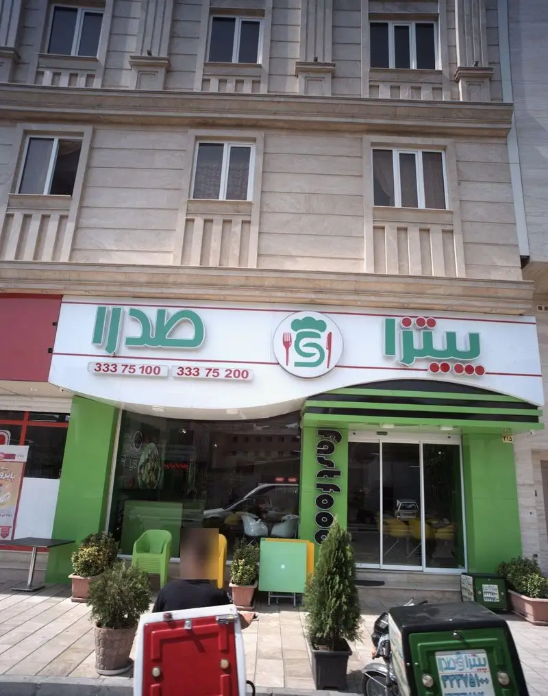 نمای بیرونی و ورودی اصلی فست فود صدرا با تابلوی چشمگیر سبز و سفید، شماره تلفن‌های تماس، لوگوی غذاخوری و پیک موتوری آماده سرویس‌دهی.