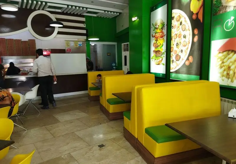 فضای داخلی فست فود صدرا با دکوراسیون مدرن سبز و زرد، شامل غرفه‌های راحت، پیشخوان سفارش، مشتریان و پوسترهای غذای لذیذ مانند پیتزا و برگر.