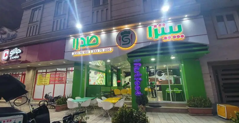 نمای بیرونی فست فود و پیتزا صدرا در شب، با تابلوهای سبز و سفید روشن و میز و صندلی‌های فضای باز.