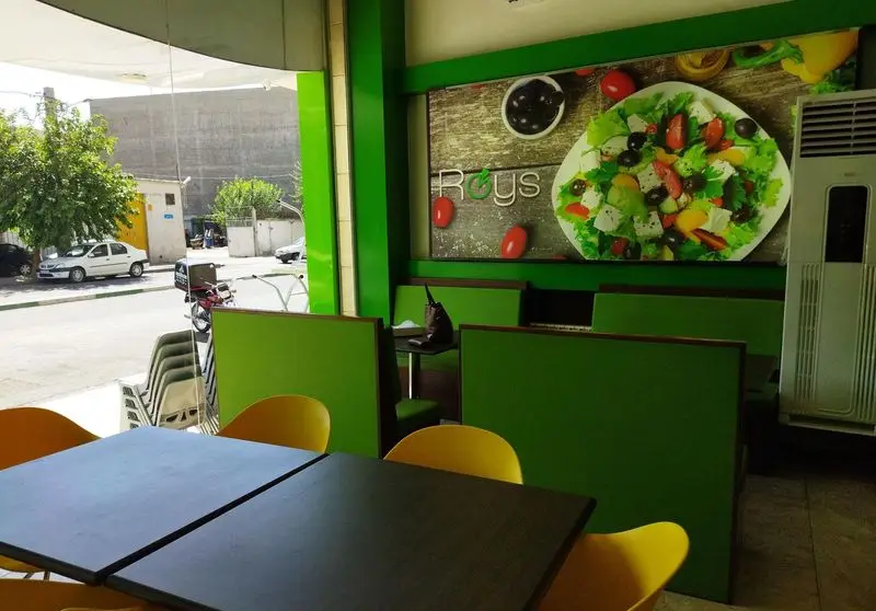 تصویری از فضای داخلی دلنشین فست فود صدرا، با دکوراسیون مدرن سبز و زرد، میز و صندلی‌های ناهارخوری و پوستر بزرگ سالاد.