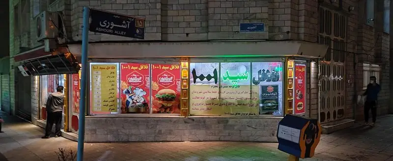 نمایی از فست فود فلافل سید ۱۰۰۱ در شب با ویترین نورانی، نمایش منوی فلافل و ساندویچ‌های متنوع، واقع در کوچه عاشوری.