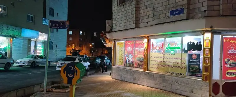 نمای شبانه فلافل سید ۱۰۰۱ در کوچه آشوری تهران، با تابلوهای روشن و ساندویچ‌ها در کنار مشتریان و خودروها.