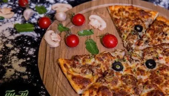 پیتزا مخصوص خوشمزه نیک برگر با تکه‌های مرغ، قارچ تازه، زیتون سیاه و گوجه گیلاسی در پس زمینه آردپاشی شده، مناسب سفارش از نیک برگر.