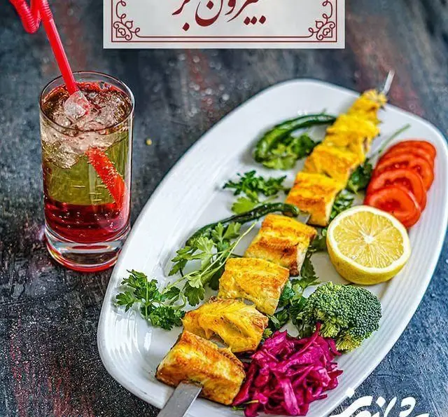 تصویر جذاب کباب ماهی مخصوص رستوران شاندیز، سرو شده با سبزیجات تازه، گوجه، لیمو، کلم قرمز و نوشیدنی خنک. انتخابی سالم و لذیذ.