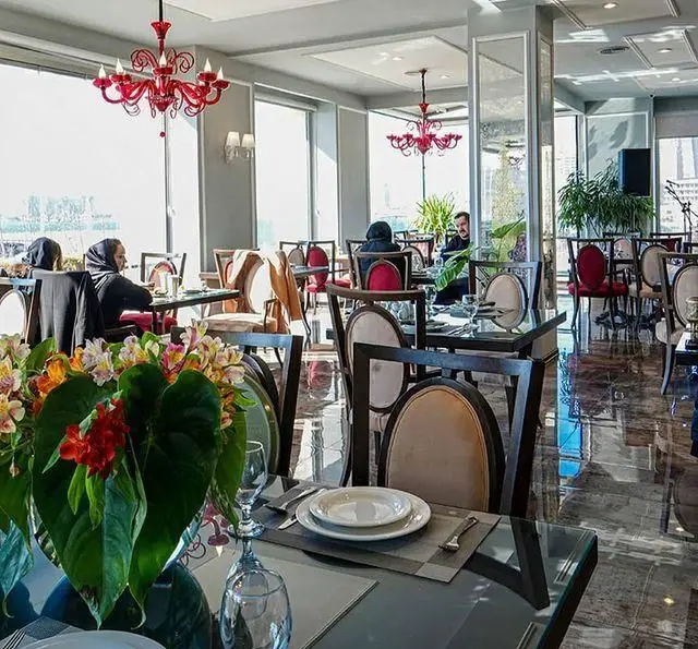 نمایی زیبا از فضای داخلی رستوران شاندیز با دکوراسیون شیک، میزهای چیده شده، لوسترهای قرمز و مشتریان در حال صرف غذا.