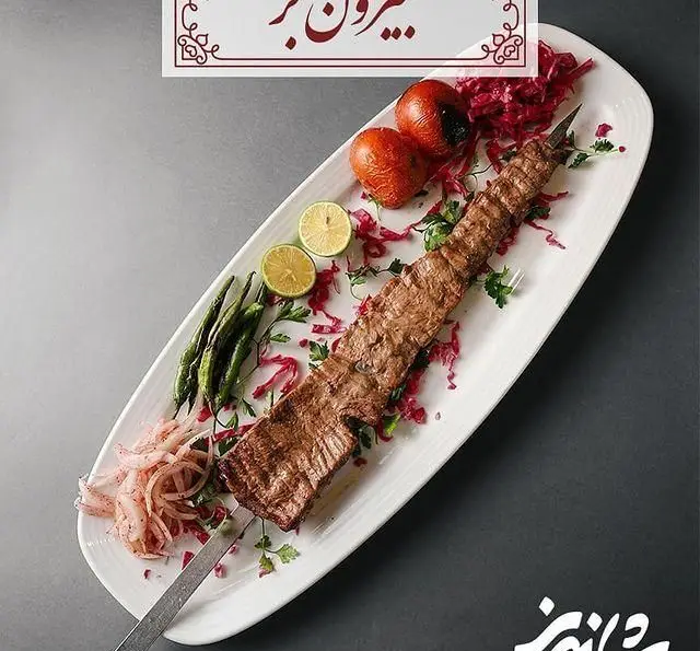 کباب برگ بلند و خوش طعم رستوران شاندیز، با تزئین گوجه فرنگی کبابی، فلفل سبز، لیموترش و پیاز قرمز روی بشقاب سفید.