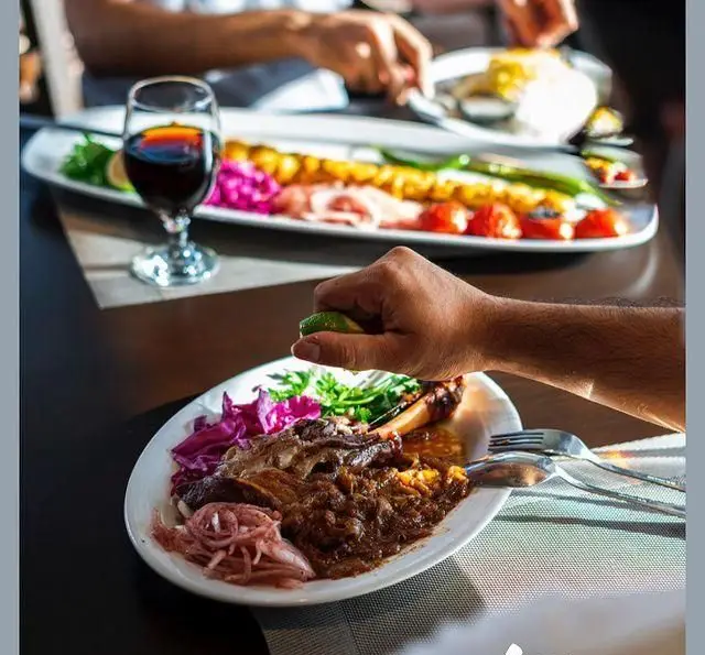 دستی در حال فشردن لیمو بر روی خورش خوشمزه در رستوران شاندیز. در پس‌زمینه انواع کباب، گوجه و فلفل کبابی دیده می‌شود.