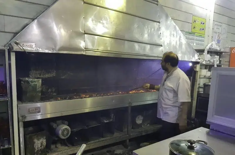 آشپز کبابی غدیر در حال آماده سازی و پخت کباب های خوش طعم و تازه روی منقل زغالی بزرگ با هود استیل صنعتی.