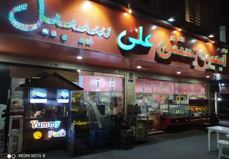 نمای بیرونی آبمیوه بستنی علی سیبیل در شب با تابلوهای روشن و غرفه بیرونی یامی پک، انواع نوشیدنی و میوه.