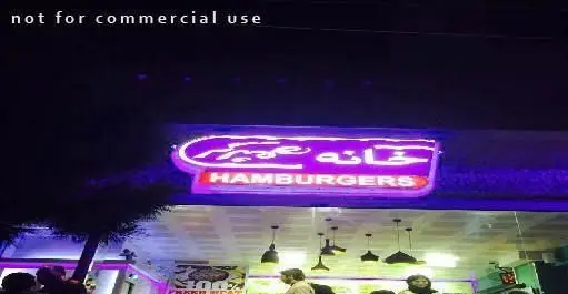 نمای خارجی "خانه همبرگر" در شب با تابلوی نئون بنفش رنگ که نام رستوران را به فارسی و انگلیسی نشان می‌دهد، همراه با مشتریان و نورهای داخلی.