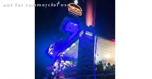 ورودی رستوران خانه همبرگر در شب، با تابلوی نئون همبرگر و نام تجاری، نورپردازی آبی چشم‌نواز و مردم در حال لذت بردن از فضای فست‌فود