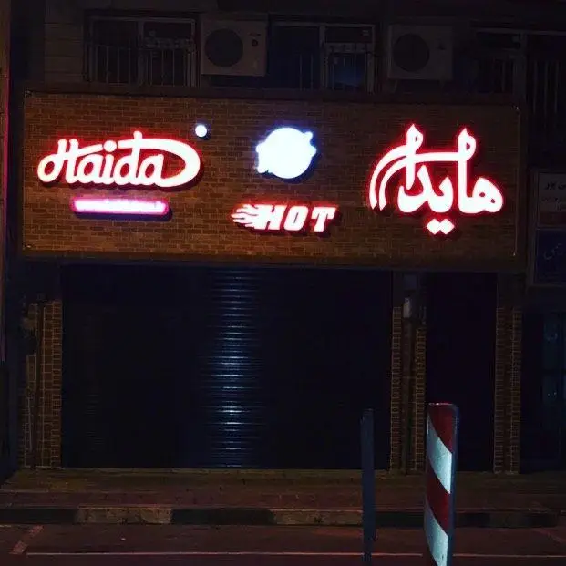 تابلو روشن فست فود هایدا هات با حروف Haida و هایدا به همراه لوگوی شعله و کلمه HOT، در نمای شب از فروشگاهی با کرکره بسته.