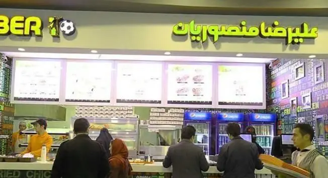 نمایی از فست فود علیرضا منصوریان، رستورانی مدرن با منوی دیجیتال و پیشخوان شلوغ. مشتریان در حال سفارش انواع غذاهای فست فود. برندینگ BER 10.