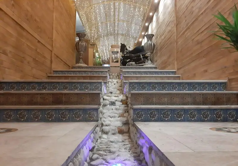 دکوراسیون داخلی لوکس کافه رستوران آرشا با پله‌های کاشی‌کاری، آبنمای مرکزی، سقف ستاره‌ای و موتورسیکلت دکوری در ورودی.