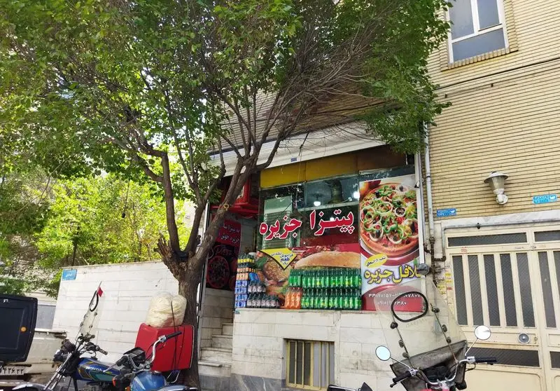 نمای بیرونی مغازه فلافل جزیره و پیتزا جزیره، با تابلوی بزرگ پیتزا، ساندویچ و نوشیدنی‌ها، همراه با درخت و موتورسیکلت‌های مشتریان در جلو.