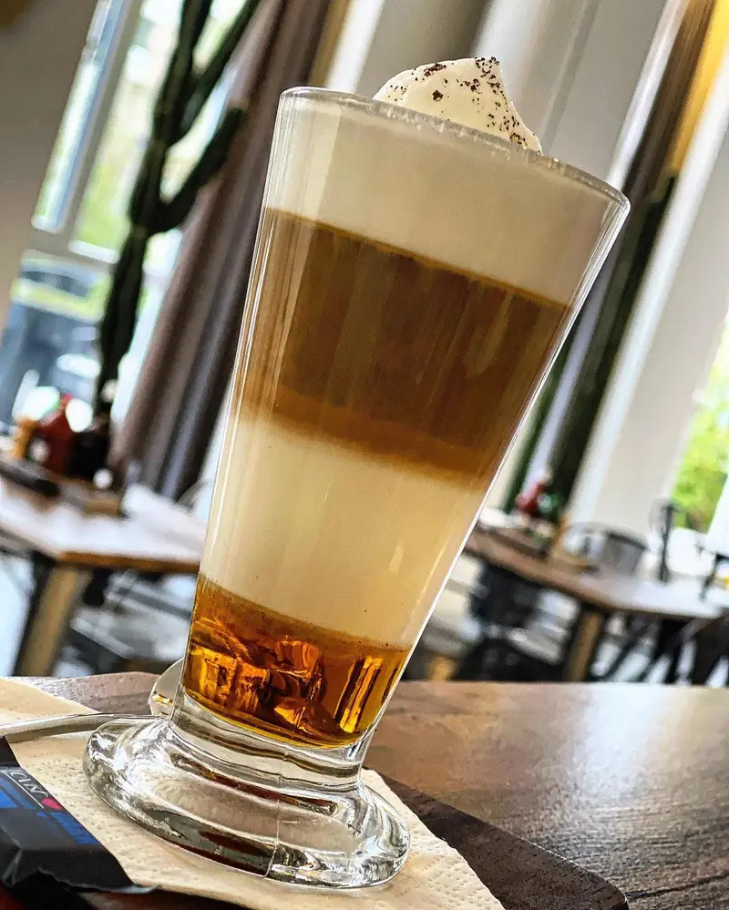 نوشیدنی قهوه لایه ای زیبا و خوشمزه در بل کافه، با خامه و پودر کاکائو روی آن، آماده سرو در محیطی دلنشین و آرامش بخش.