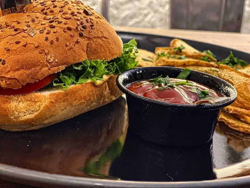 یک برگر لذیذ بل کافه با نان کنجدی، گوجه و کاهو، همراه با سیب‌زمینی تنوری طلایی و سس کچاپ، آماده سرو در محیطی دلنشین.