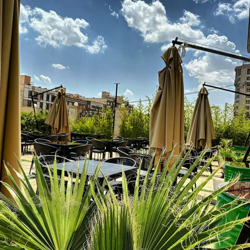 فضای باز دلنشین و آفتابی بل کافه با میز و صندلی‌های راحت، چترهای بزرگ و گیاهان سرسبز، مکانی آرام و ایده‌آل برای دورهمی.