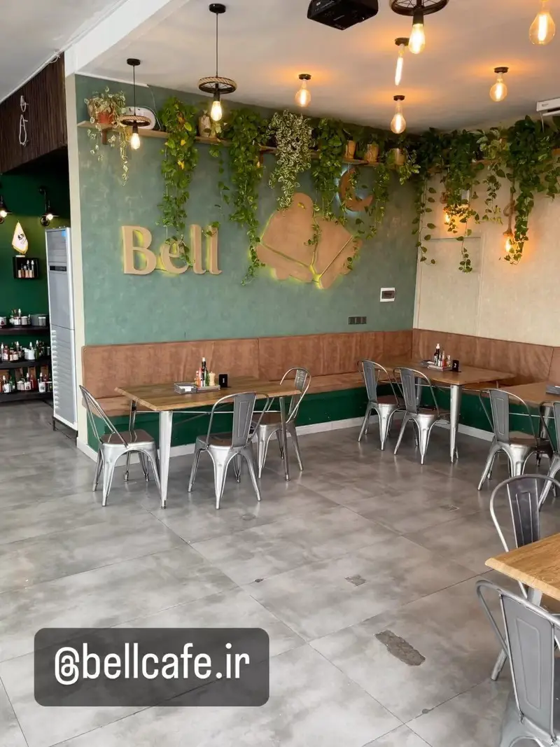 تصویر فضای داخلی بل کافه با دیوارهای سبز و چیدمان گیاهان آویز، مبلمان چوبی و صندلی‌های مدرن فلزی، فضایی دنج و دلنشین با نورپردازی گرم.
