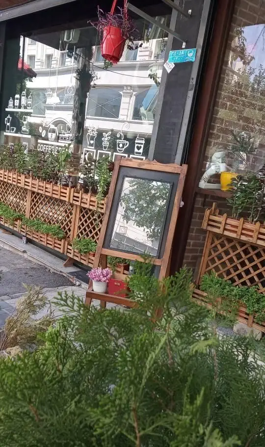 نمای بیرونی کافه رستوران جوان با پنجره‌های طرح‌دار قهوه، نرده چوبی سبز و آینه دکوری روی سه‌پایه.