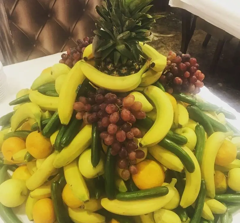 نمایش باشکوه میوه و سبزیجات تازه شامل موز، انگور، خیار، پرتقال و آناناس در بوفه رستوران جوان هنرور ۲.