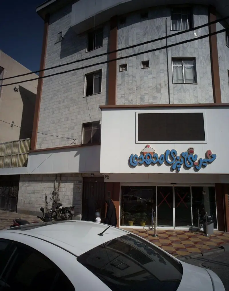 نمای بیرونی شیرینی فروشی محمودی با تابلوی آبی رنگ و نماد کاپ کیک در ساختمان سنگی، همراه با خودرویی در پیش زمینه خیابان.