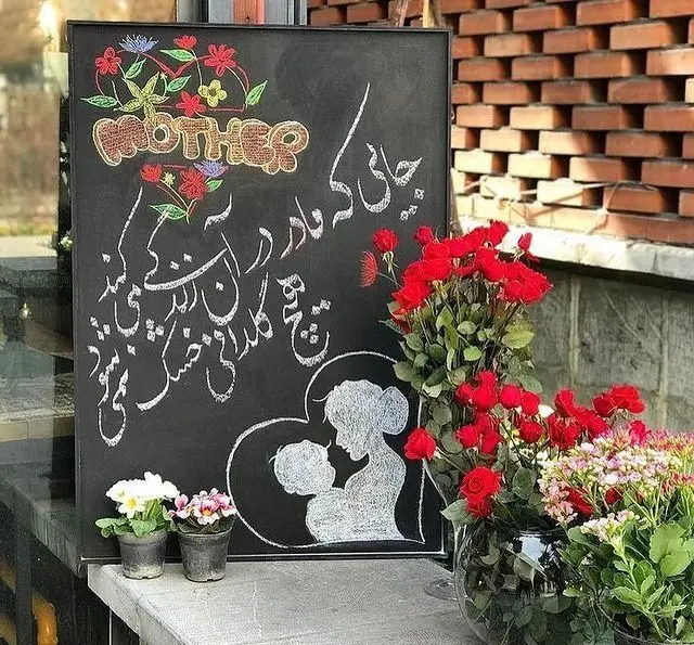 تخته سیاه کافه مونو با طراحی مادر و فرزند، شعر فارسی و کلمه Mother، تزئین شده با گل‌های رز قرمز فراوان و گلدان‌های کوچک، مناسب روز مادر.