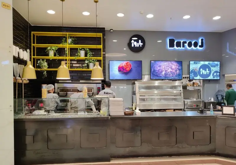 نمای داخلی رستوران باروژ با پیشخوان مدرن، آشپزخانه باز، کارکنان در حال کار، و نمایشگرهای منوی غذا و دکوراسیون شیک.