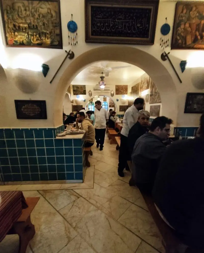 فضای گرم و سنتی رستوران دیزی سرا با معماری طاق‌دار، تابلوهای هنری و کاشی‌کاری آبی، مشتریان در حال صرف دیزی ایرانی.