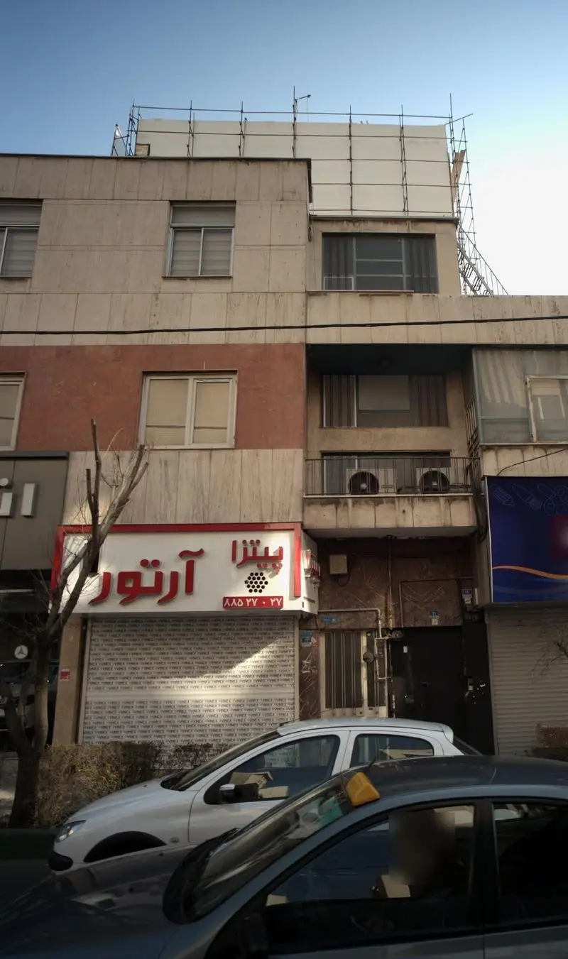 تابلوی قرمز و سفید فست فود پیتزا آرتور با شماره تلفن 88577-27 در نمای بیرونی مغازه، همراه با کرکره بسته و خودروها در خیابان تهران.